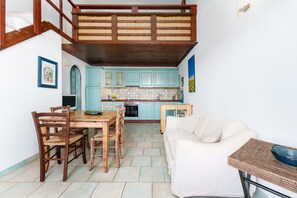 A dining area with a wooden table and chairs, a white sofa, a kitchen with light blue cabinets, and a staircase. - Orosei Domo Holiday Home with Mountain View, Private Terrace & Air Conditioning (Orosei)