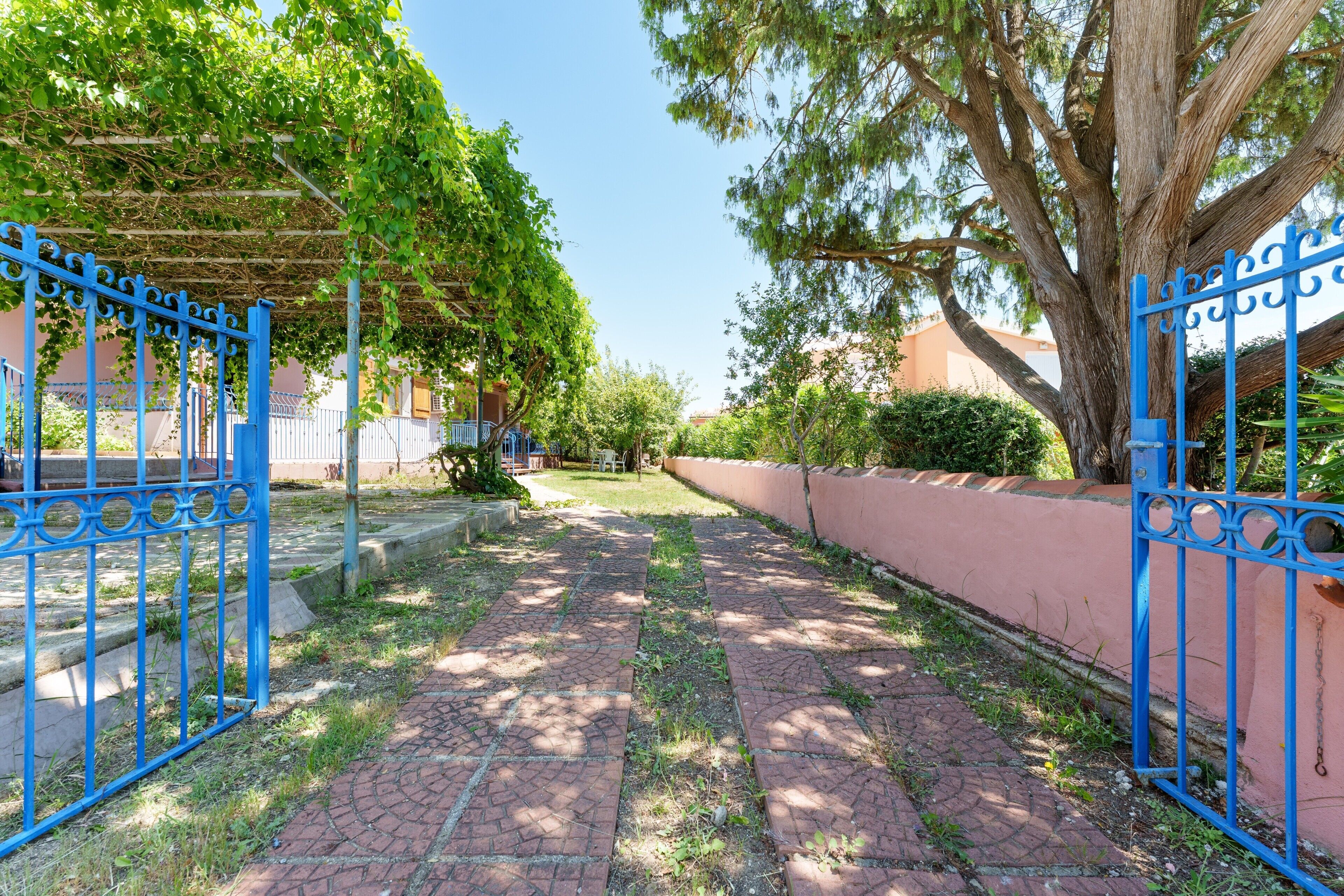 A paved pathway leading to a blue gate, surrounded by greenery and a building with a pink wall.