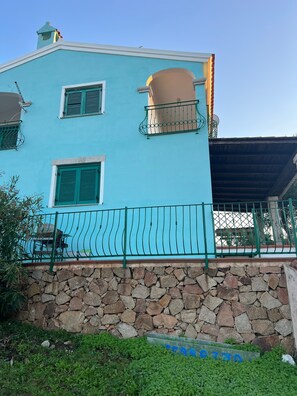 Ein zweistöckiges Gebäude mit einer steinernen Fundamentwand, einem Balkon mit Metallgeländer und blauem, wolkenlosem Himmel.