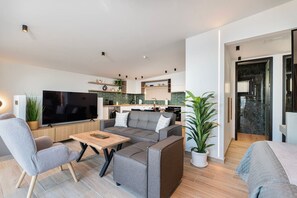 Ein modernes Wohnzimmer mit einem grauen Sofa, einem Holztisch und einem Flachbildfernseher. Im Hintergrund ist eine Küche mit grüner Rückwand und weißen Schränken zu sehen.