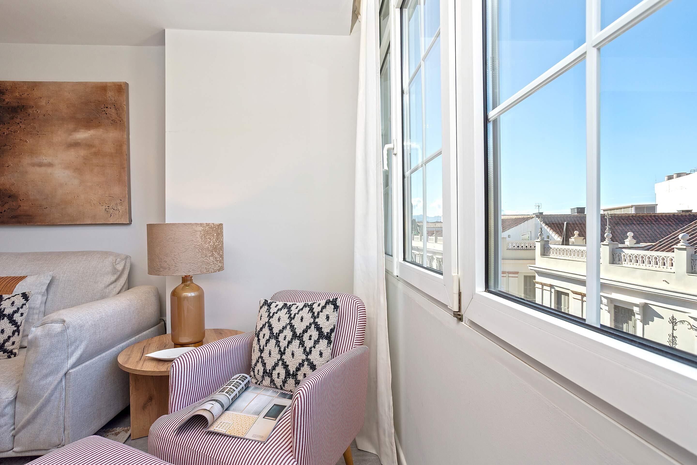 Una habitación de esquina con un ventanal grande, un sofá, una silla y una mesita con una lámpara.