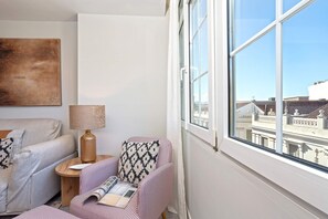 Ein Eckzimmer mit großem Fenster, einer Couch, einem Sessel und einem kleinen Tisch mit Lampe.