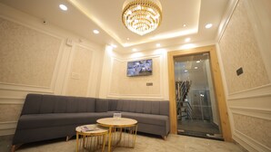 A modern lobby with a grey sofa, two round tables, a chandelier, and a TV mounted on the wall. - BABA PALACE (Ayodhya)