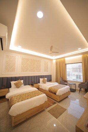 A hotel room with two beds, a ceiling fan, a window with curtains, and a small table. - BABA PALACE (Ayodhya)