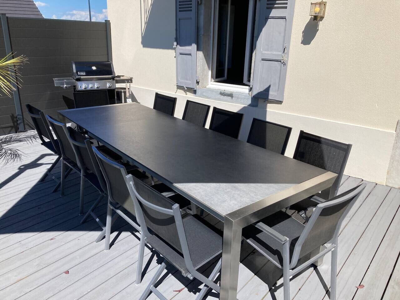 Un espace de restauration en plein air avec une longue table et des chaises, un barbecue et une fenêtre avec des volets.