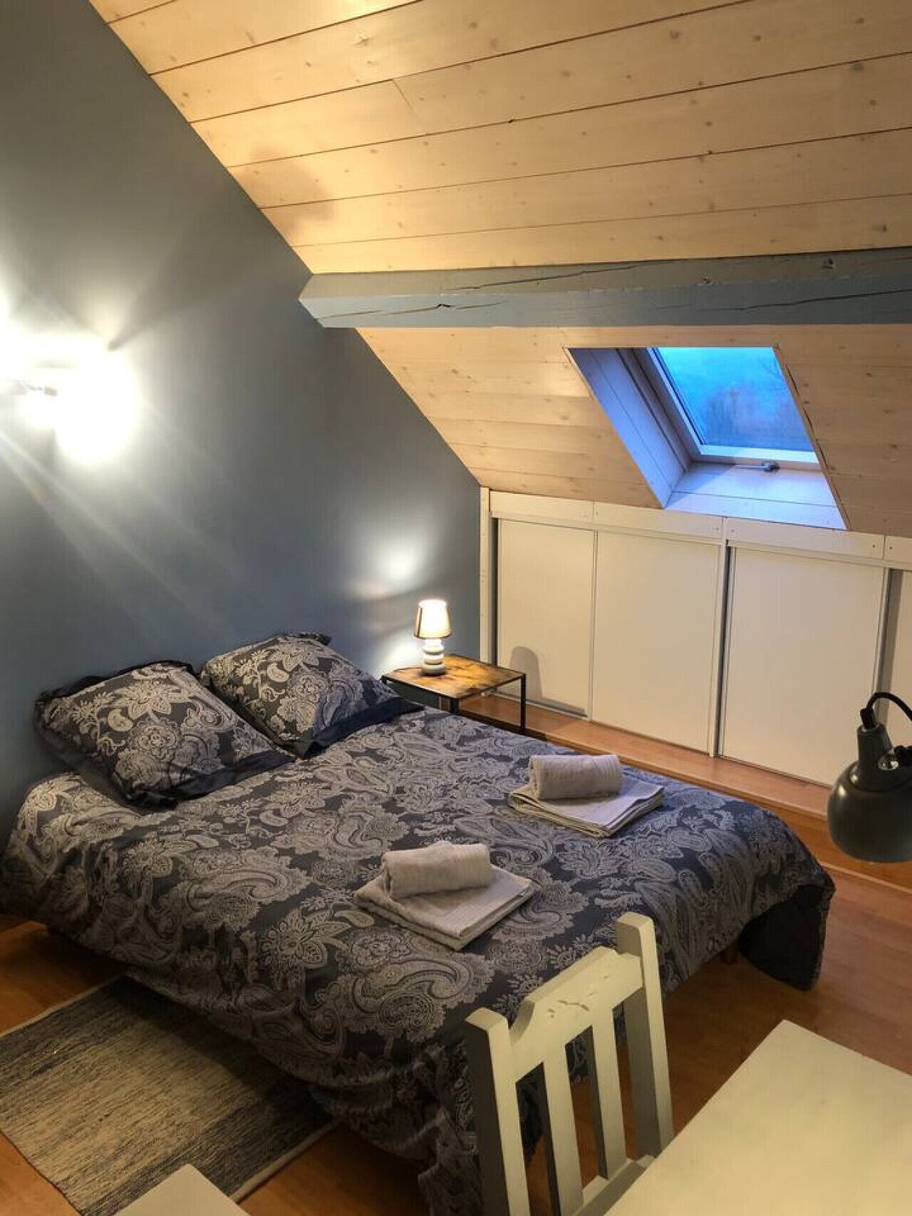 Une chambre avec un plafond en pente, un puits de lumière, un lit avec une literie à motifs, une table de chevet avec une lampe et une chaise.