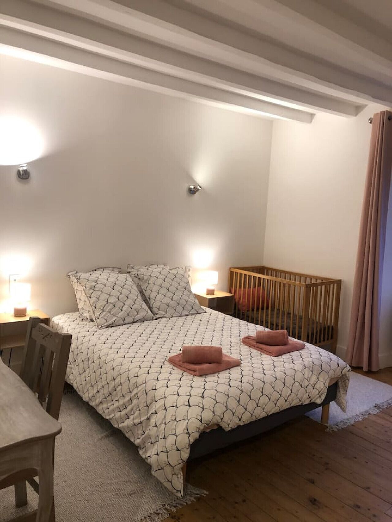 A bedroom with a bed, bedside tables, a wooden crib, and a chair.