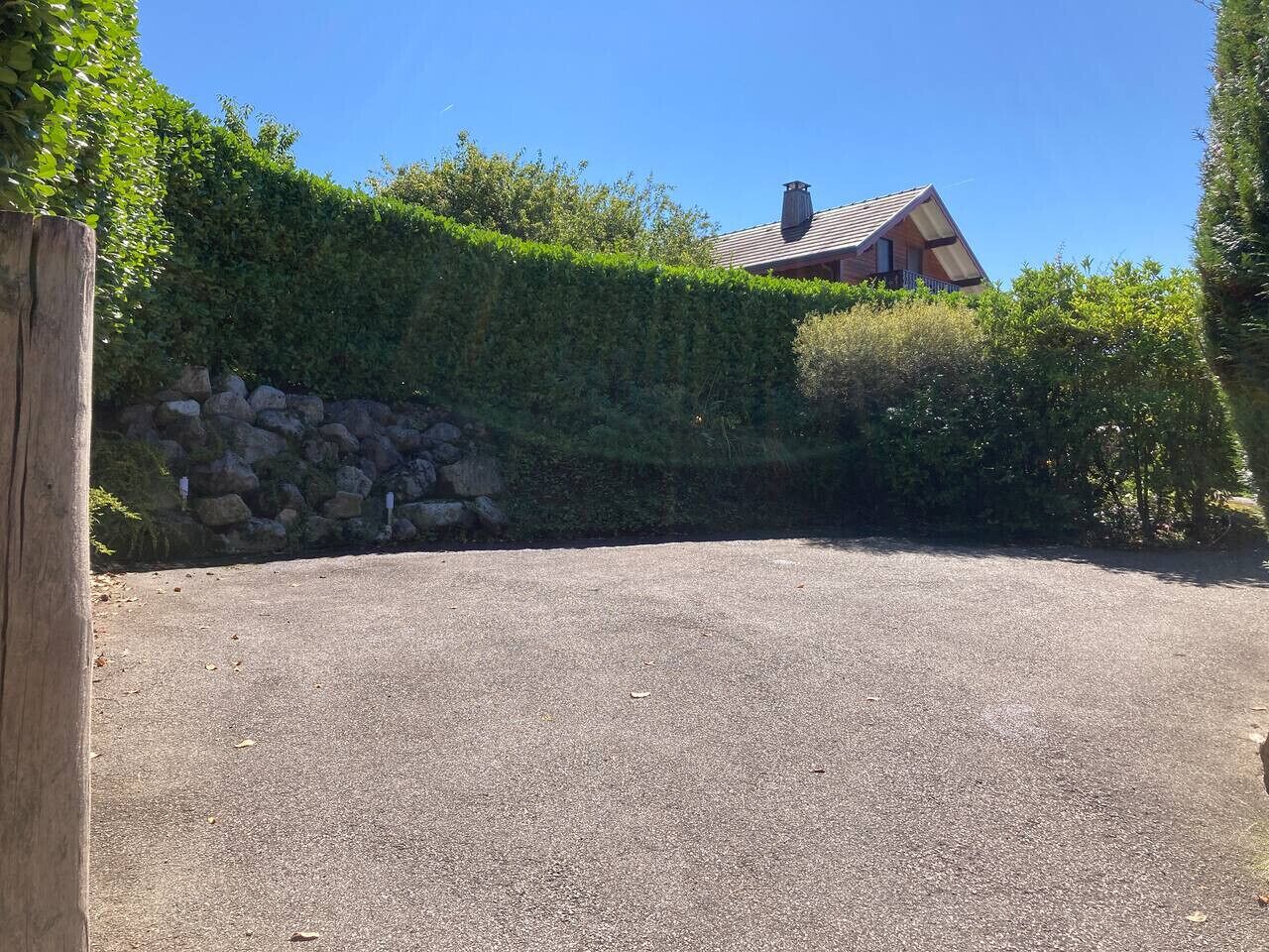 A gravel driveway leading to a house with a sloped roof, surrounded by dense green hedges and a stone wall.
