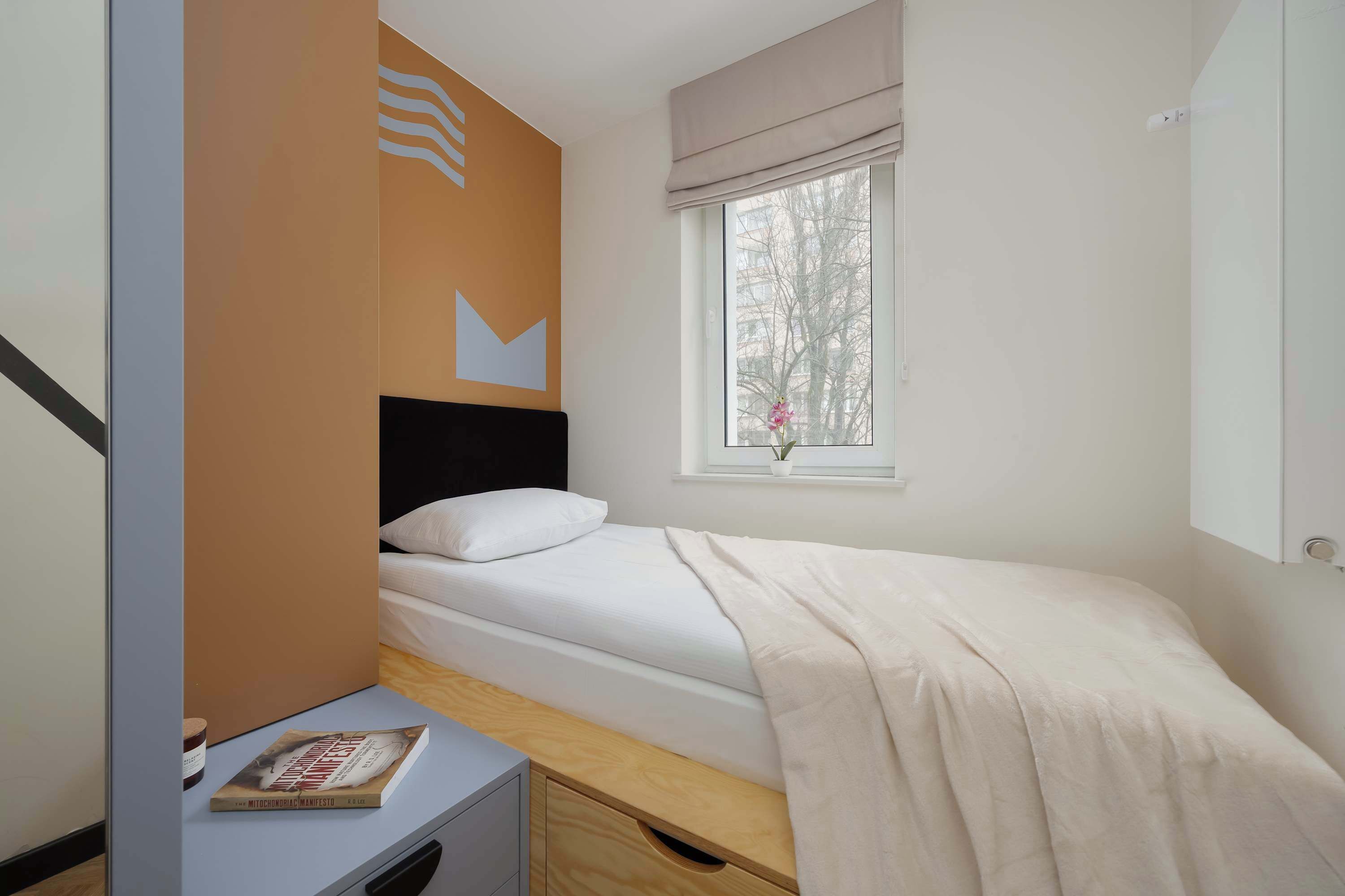 A bedroom with a single bed, a bedside table with a book, a window with a view of trees, and a wall with a geometric design.