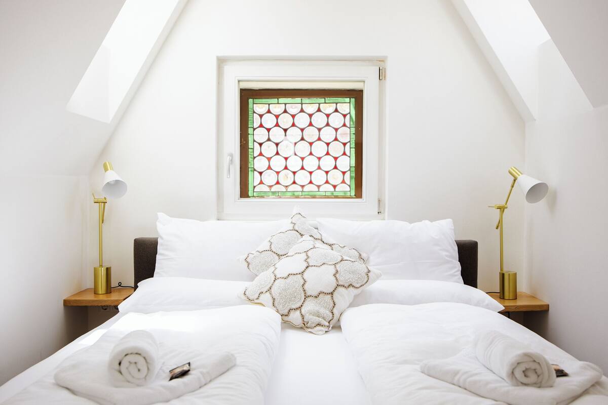 Een keurig opgemaakt bed met witte lakens en een decoratief kussen, twee bedlampjes en een raam met een geometrisch patroon.