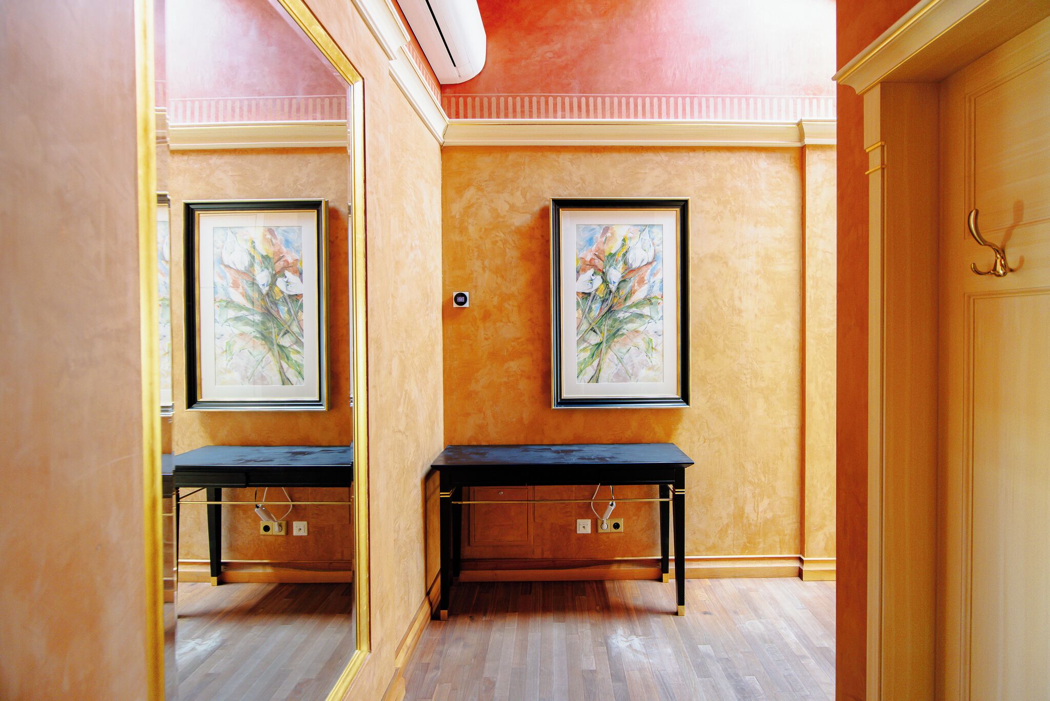 A hallway with two framed paintings on the walls, a black bench, and a wooden door with a gold handle.