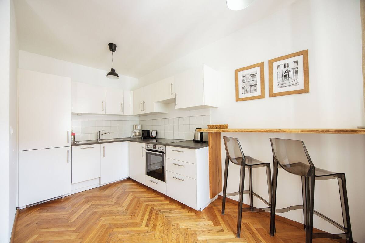 Een moderne keuken met witte kasten, een houten visgraatvloer, een inbouwoven en twee barkrukken.