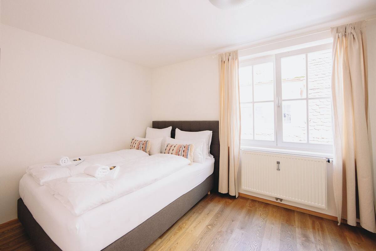 Een net opgemaakt bed met witte lakens en handdoeken, een donker hoofdbord en een raam met gordijnen.