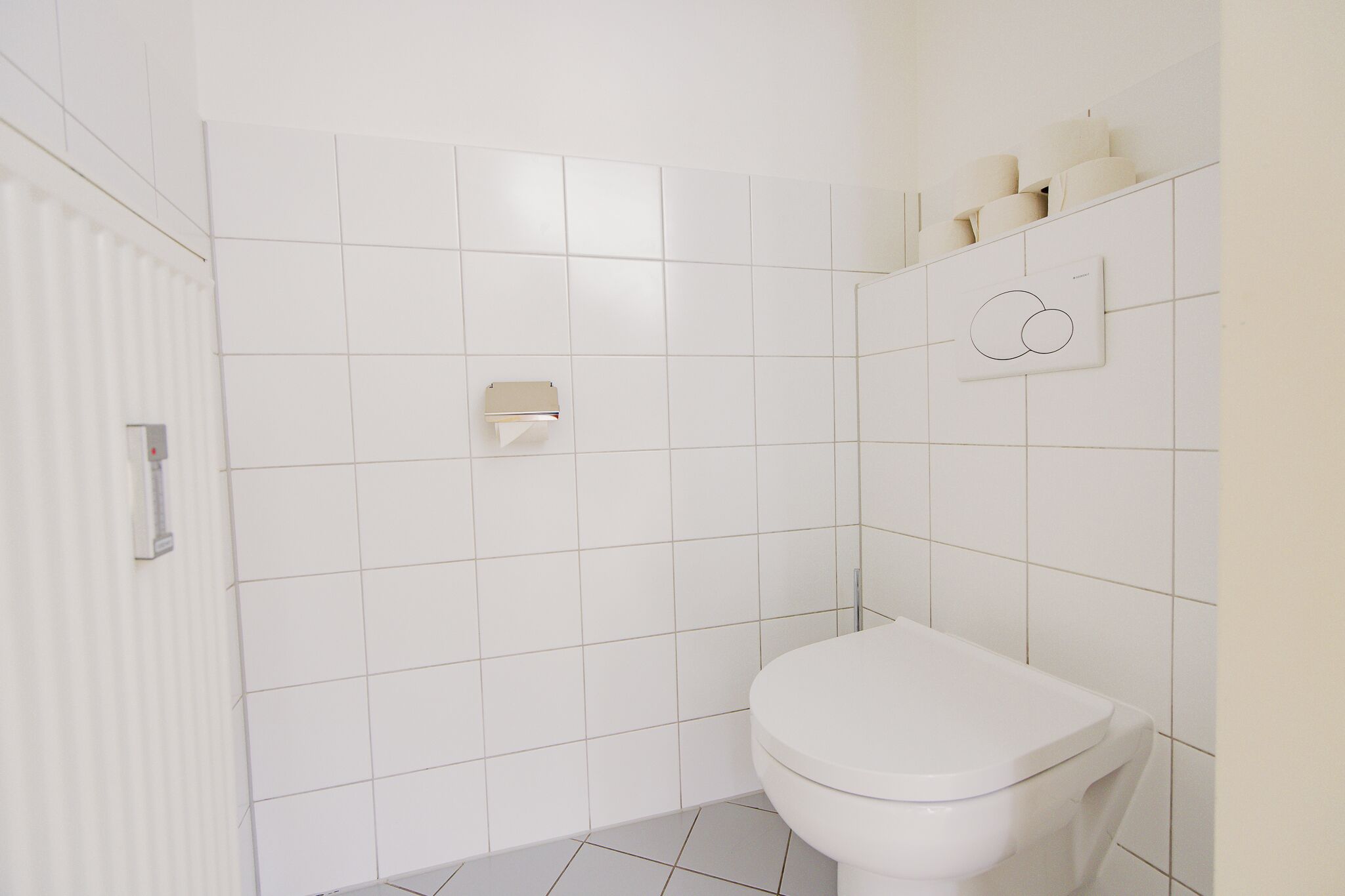 Ein Badezimmer mit weißen Fliesen, einer Toilette, einem Handtuchspender und einem an der Wand befestigten Toilettenpapierhalter.
