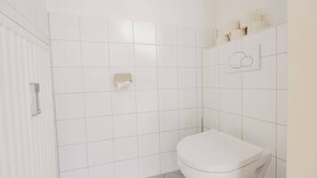 Ein Badezimmer mit weißen Fliesen, einer Toilette, einem Handtuchspender und einem an der Wand befestigten Toilettenpapierhalter.