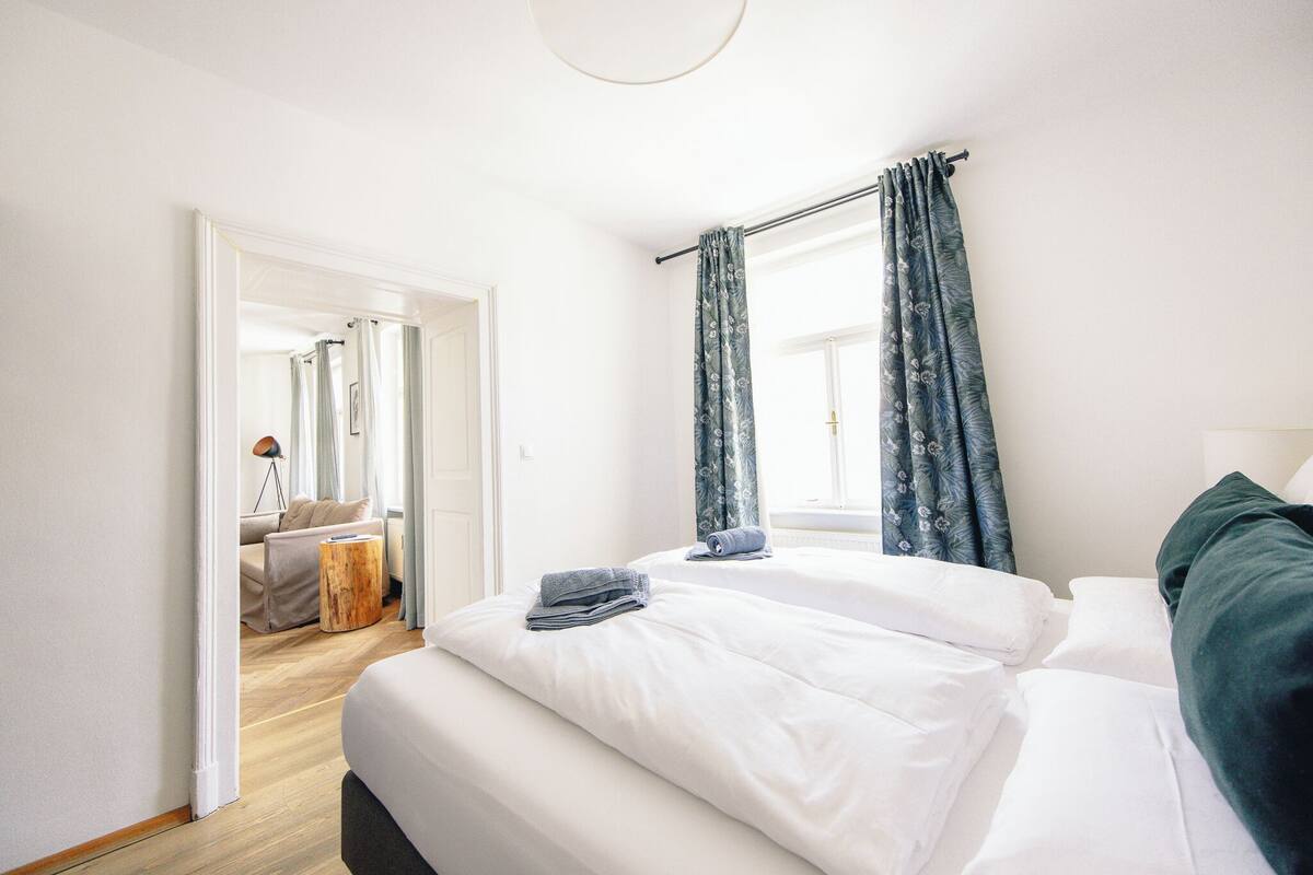 Een hotelkamer met een groot bed, een klein houten nachtkastje en uitzicht op de gang.