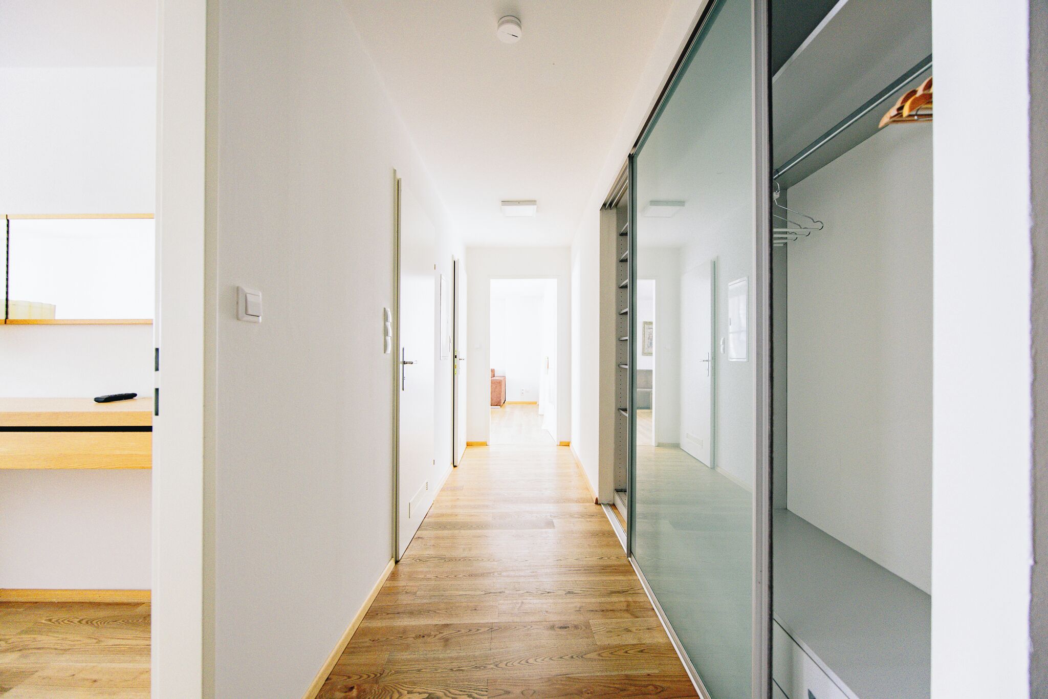 A hallway with wooden flooring, white walls, and a glass partition on the right.