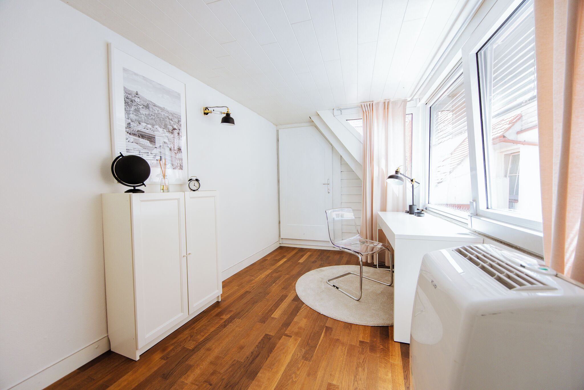 Une pièce avec un plancher en bois, des murs blancs, un bureau avec une chaise, un radiateur et une fenêtre avec des rideaux.