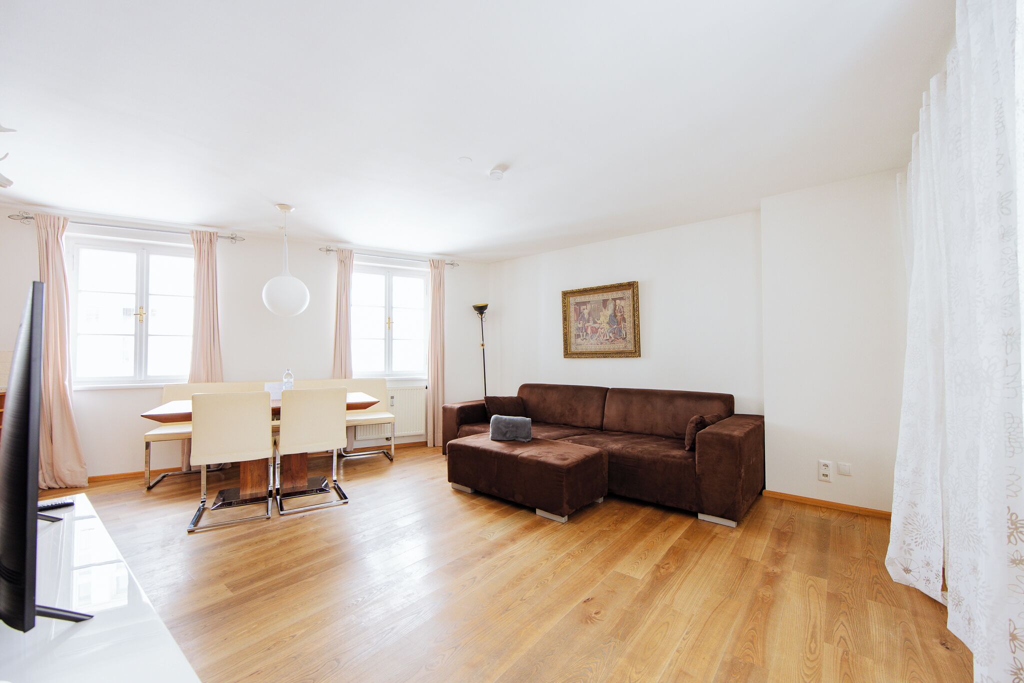 A modern living room with a brown sectional sofa, a dining table with chairs, a flat-screen TV, and a painting on the wall.