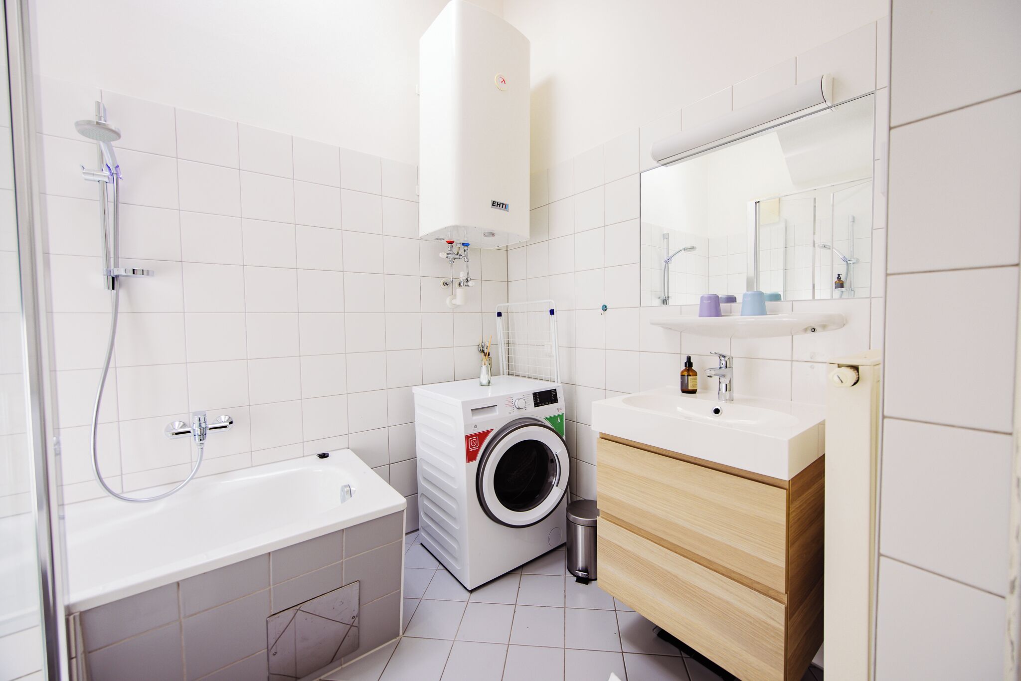 A modern bathroom with a white tiled floor, a white bathtub, a washing machine, a sink with a mirror above it, and a wooden cabinet.