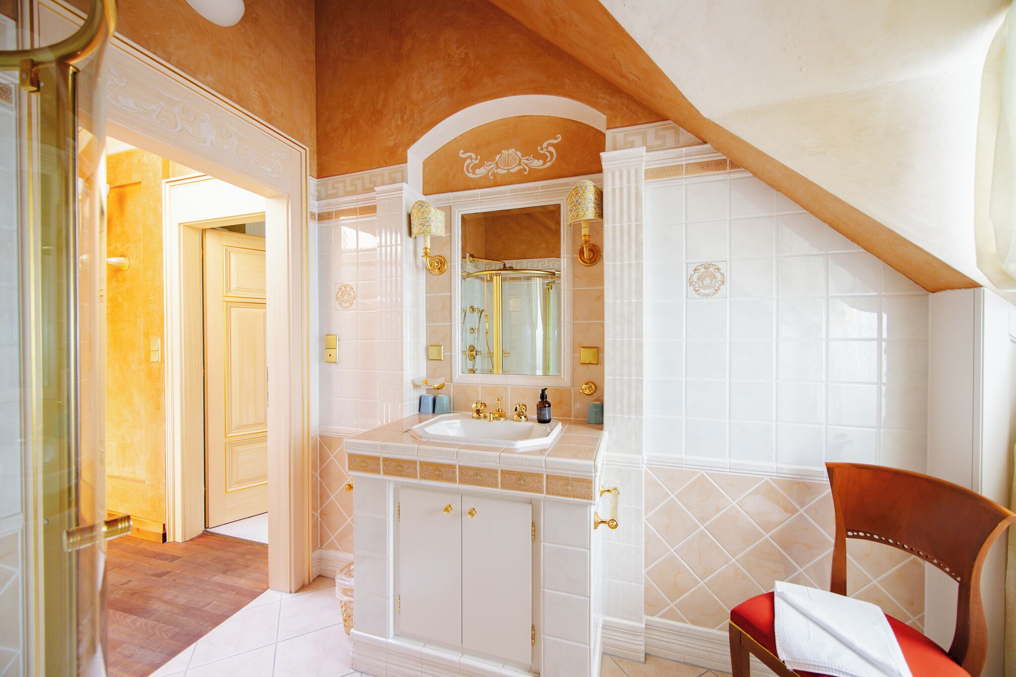 Un baño con tocador blanco, detalles dorados y una silla de madera.