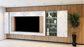Ein modernes Wohnzimmer mit einem großen Flachbildfernseher, der an einer Marmorwand angebracht ist, Holzwandverkleidung und einer Einbauschränke mit Dekorationsobjekten.