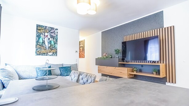 Una sala moderna con un televisor de pantalla plana grande montado en una pared de paneles de madera, un sofá gris con cojines azules y una mesa de centro con un adorno.