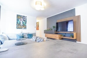 Ein modernes Wohnzimmer mit einem großen Flachbildfernseher an einer Holzpaneelwand, einem grauen Sofa mit blauen Kissen und einem Couchtisch mit einem dekorativen Gegenstand.