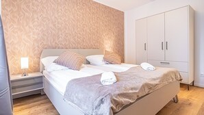 Una cama doble bien hecha con una colcha gris y dos toallas, una mesita de noche con una lámpara y un armario de dos puertas.