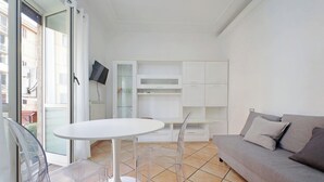 Ett modernt vardagsrum med ett runt, vitt bord, genomskinliga stolar, en grå soffa och inbyggda, vita hyllor.