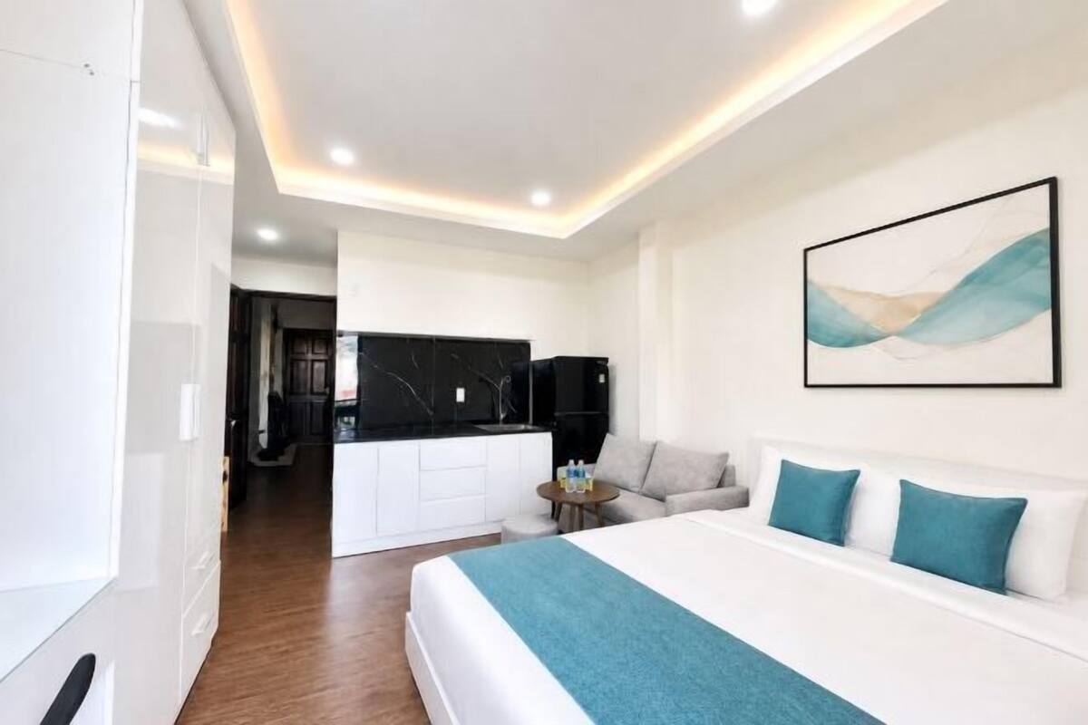 A modern hotel room with a large bed, a small seating area, a kitchen area, and a painting on the wall.
