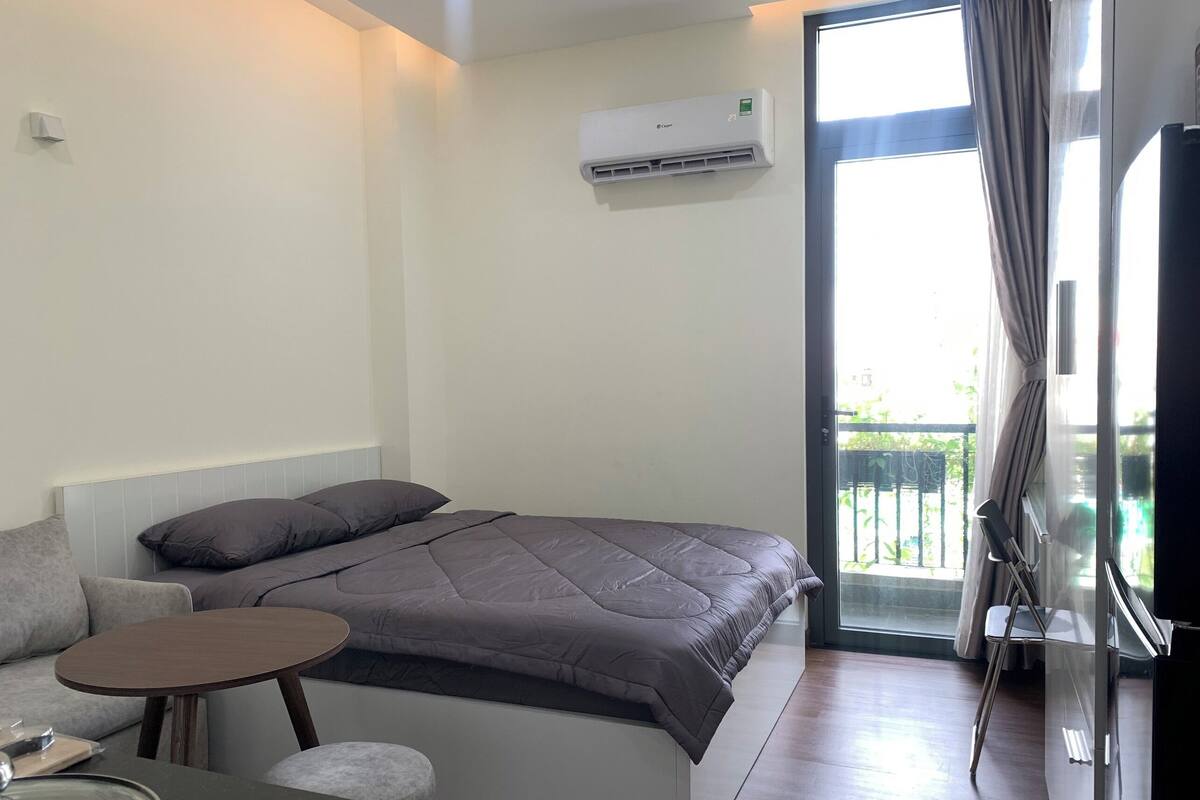 A modern bedroom with a bed, a small table, and a chair. There is a window with curtains and an air conditioning unit on the wall.