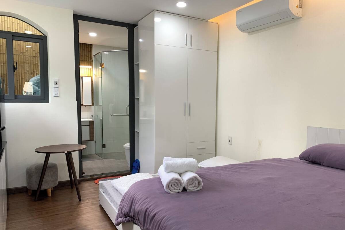 A hotel room with a bed, a small table, a wardrobe, and a bathroom visible through a glass door.