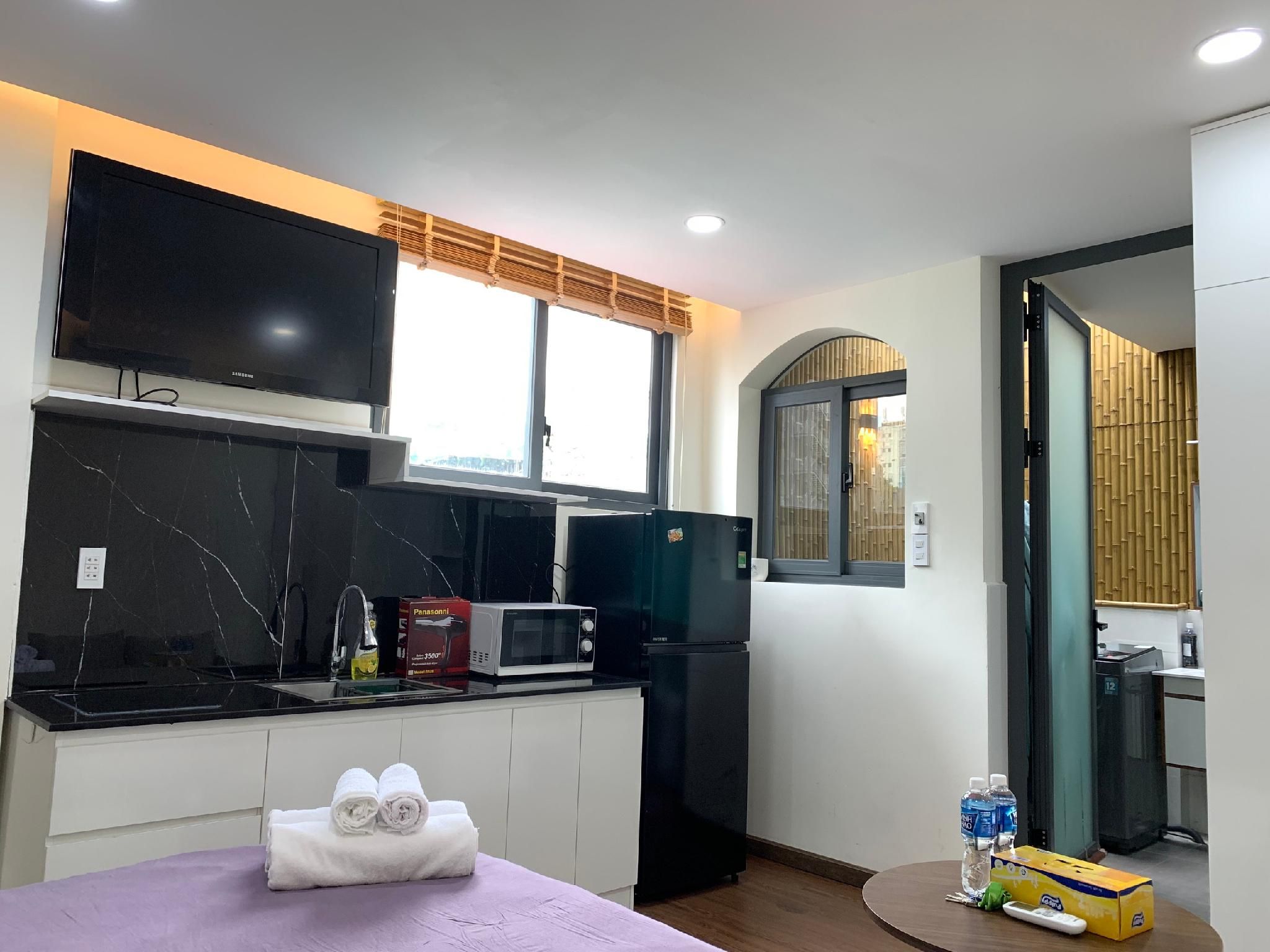 Una cocina moderna con una televisión de pantalla plana, microondas y refrigerador. Hay una ventana con persiana de bambú, y una pequeña mesa con toallas.