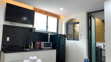 Una cocina moderna con una televisión de pantalla plana, microondas y refrigerador. Hay una ventana con persiana de bambú, y una pequeña mesa con toallas.