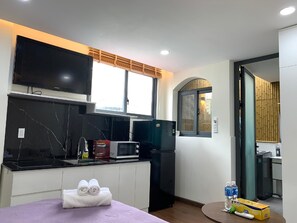 A modern kitchen with a flat-screen TV, a microwave, and a refrigerator. There is a window with a bamboo blind, and a small table with towels. - Lunaria Maison - Saigon Central Park, District 3 (Ho Chi Minh City)