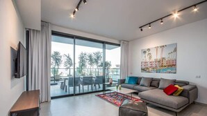 Ein modernes Wohnzimmer mit grauem Sofa, einem Couchtisch und einem Fernseher an der Wand. Ein großes Fenster mit Vorhängen, ein Gemälde an der Wand und ein Blick ins Freie.