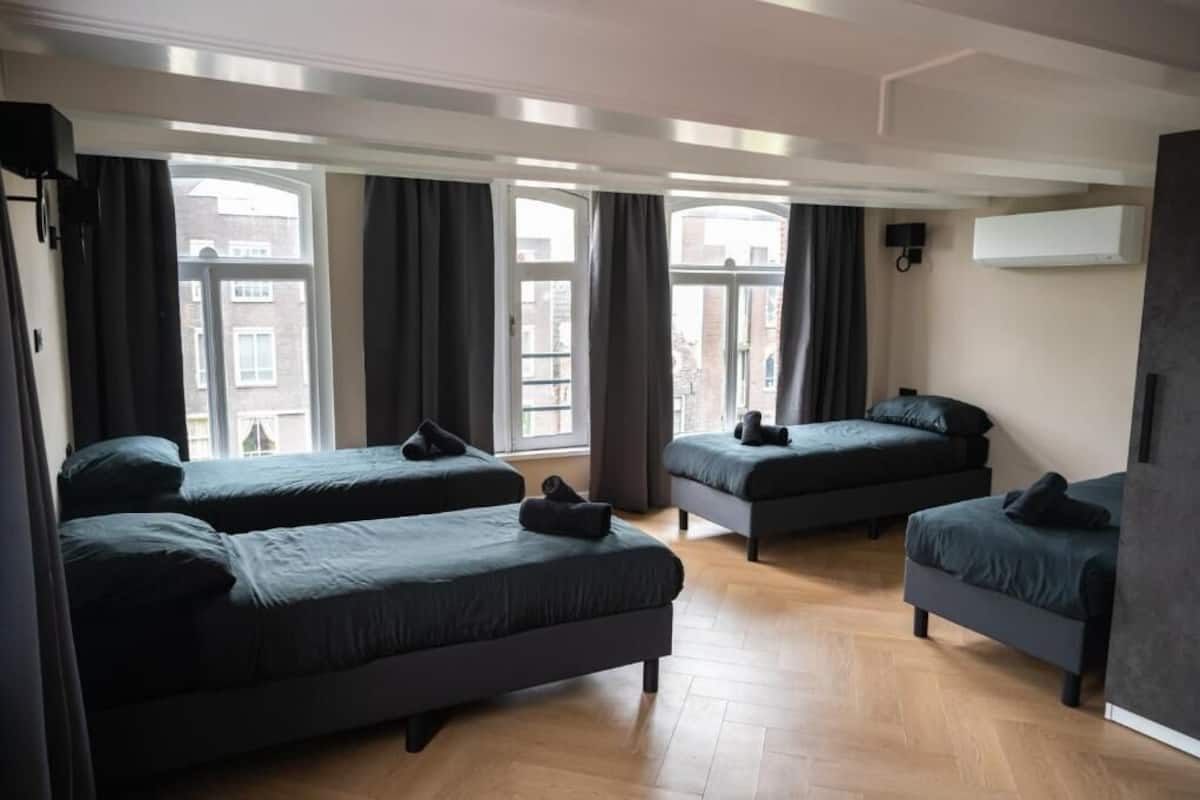 A hotel room with four single beds arranged in a row, large windows with curtains, a wooden floor, and an air conditioning unit on the wall.