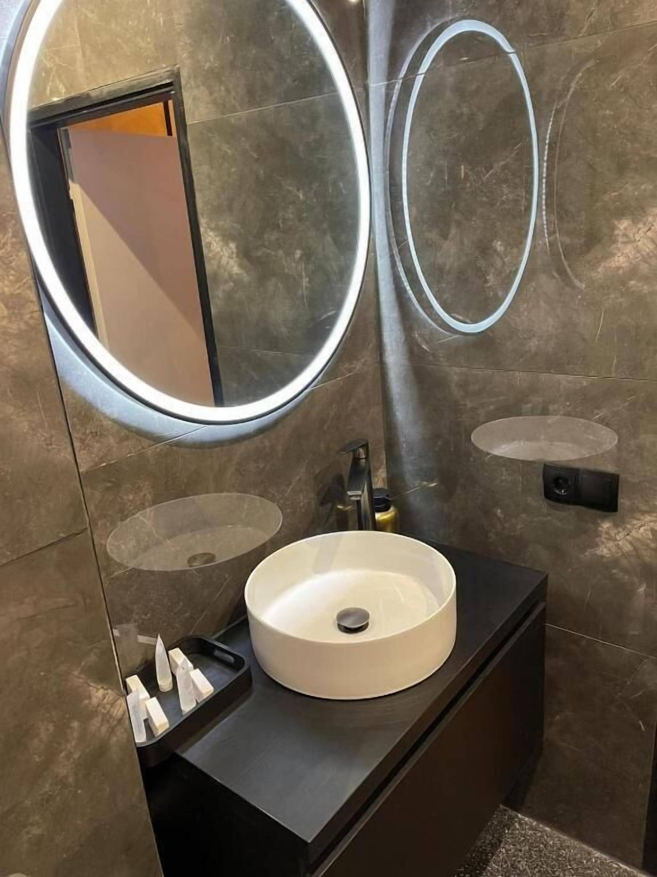 Un baño moderno con un espejo redondo, un lavabo redondo y un mueble de tocador oscuro.