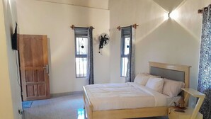 A bedroom with a wooden bed, a bedside table, a wall-mounted fan, and two windows with patterned curtains. - Gray Hideaway Resort (Kayunga)