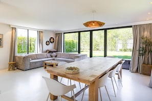 Una sala de estar moderna con una gran mesa de comedor, un sofá modular y ventanas amplias que ofrecen vistas a la vegetación.