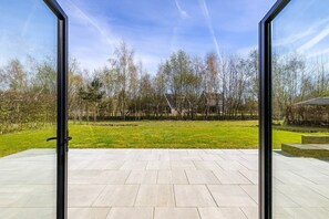 A view through glass doors onto a patio with a tiled floor, surrounded by trees and a grassy area.