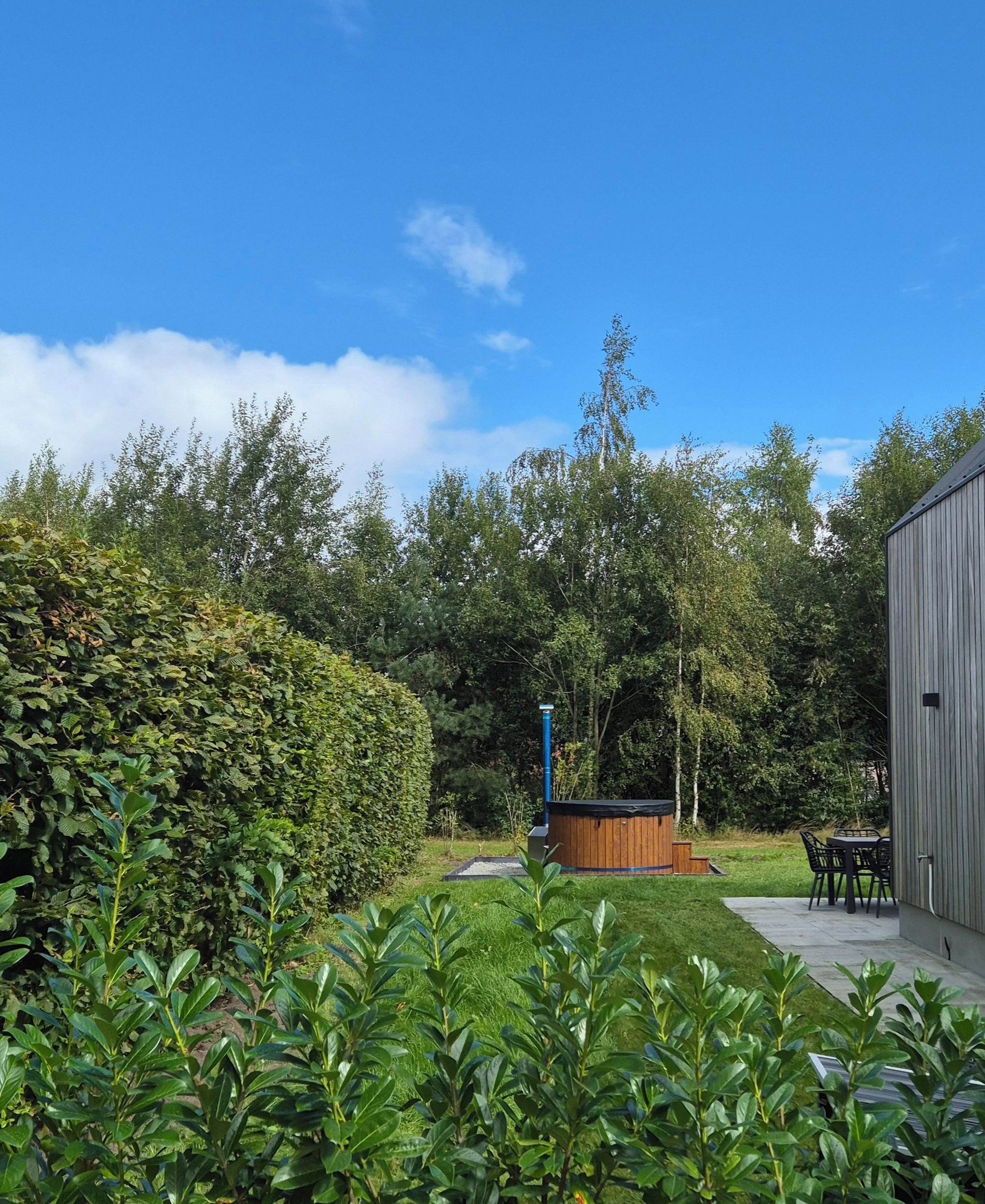 A well-maintained garden with a wooden hot tub, a metal structure, and a dining area with chairs and a table.