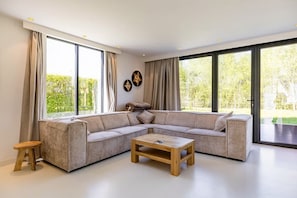 Ein modernes Wohnzimmer mit einem Ecksofa, einem Holztisch und großen Fenstern mit Blick auf Grünflächen.