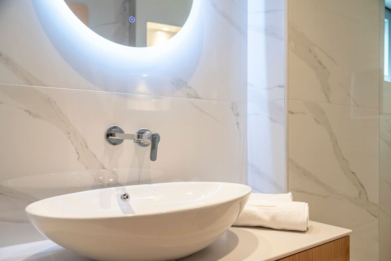 Une salle de bain avec un lavabo en marbre blanc, un robinet moderne et un miroir rond surmonté d’une lampe.
