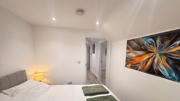 Ein Schlafzimmer mit Bett, Nachttisch mit Lampe, einem Bild an der Wand und einem Flur, der zu einem anderen Raum führt.