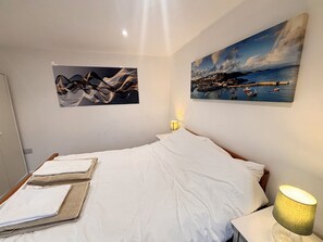 Un lit bien fait, avec du linge de lit blanc et une serviette pliée, une lampe de chevet et deux tableaux accrochés au mur représentant des scènes abstraites et des paysages côtiers.
