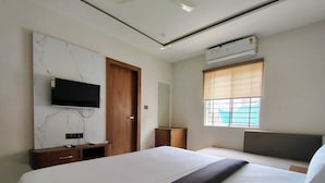 A modern hotel room with a large bed, a TV mounted on a marble wall, a wooden wardrobe, a bedside table, and a window with blinds. - Hotel Gurukripa (Ujjain)