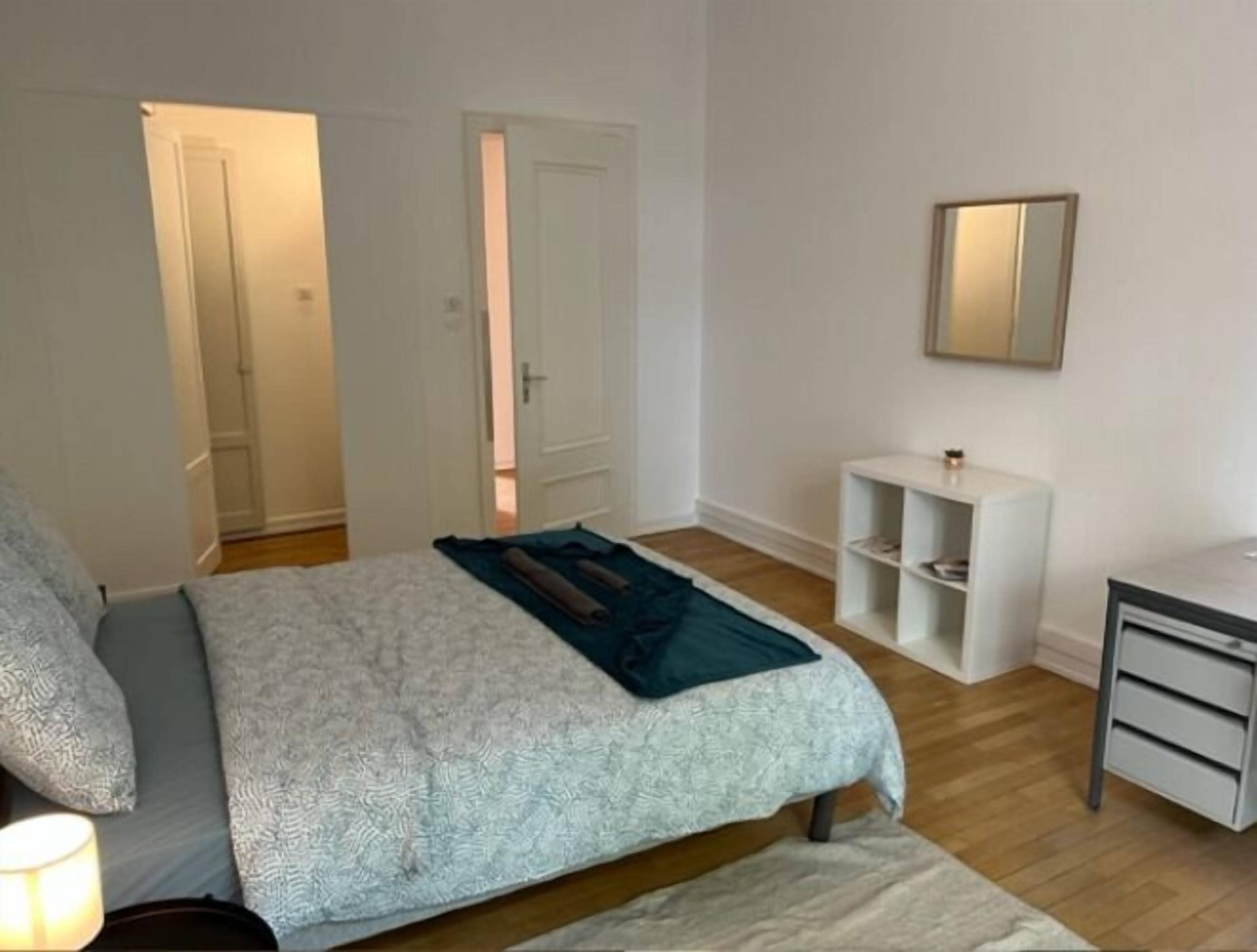 A bedroom with a bed, a bedside table, a nightstand, a mirror, and a door leading to another room.