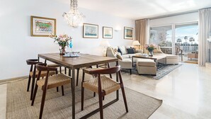 A spacious living room with a dining area, featuring a large wooden table, woven chairs, and a chandelier. There are framed artworks on the walls and a view of the outdoors through large windows. - MA - Elegant Apartment With Sea Views (Marbella)